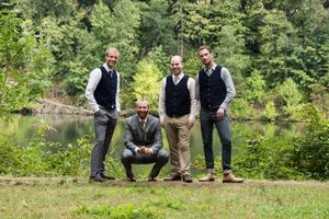 Four well-dressed men posing outdoors near a lake surrounded by lush green trees, dressed in formal attire.