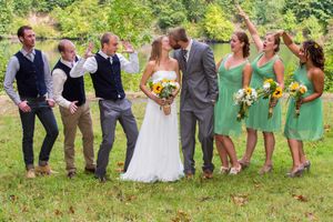 Wedding party poses outdoors, bride and groom kissing while bridesmaids in green dresses and groomsmen celebrate around them.