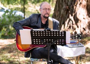 Bald man with glasses playing acoustic guitar outdoors near large tree at a woodland music event.