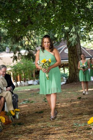 Bridesmaid in green dress walks down outdoor aisle holding sunflower bouquet at a garden wedding ceremony.