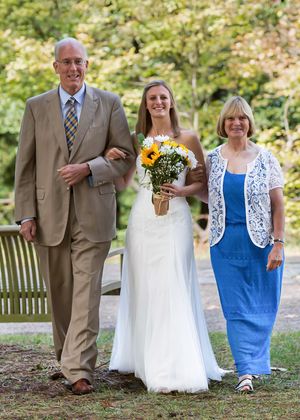 Bride in white gown holding sunflower bouquet walks with parents in formal attire at outdoor wedding ceremony.
