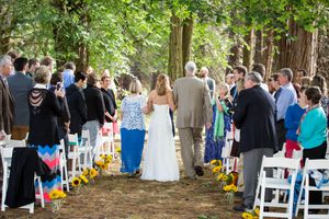 Outdoor wedding ceremony aisle lined with sunflowers, bride walking with escort through wooded venue with seated guests.
