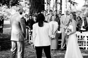 Black and white outdoor wedding ceremony with bride holding bouquet, groom, and officiant before seated guests.