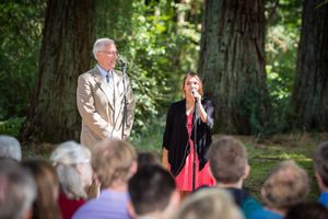 Man and woman speaking at an outdoor ceremony surrounded by tall trees with seated audience.