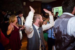 Guests dancing joyfully at a wedding reception, with a bearded man in a vest and tie raising his arms in celebration.