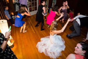 Bride in white gown dancing energetically with guests on wooden dance floor at wedding reception.
