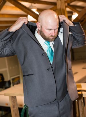 Bald bearded groom in gray suit adjusting jacket collar with teal tie at wedding venue.