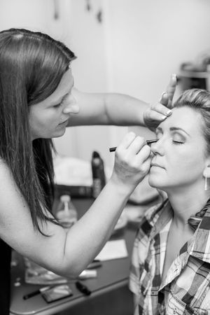 Black and white photo of a makeup artist applying eye makeup to a blonde woman in a plaid shirt.