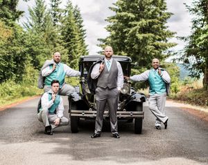 Groom and groomsmen in gray suits with teal vests posing by a vintage black car on a tree-lined road.