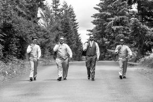 Four groomsmen walking confidently down a tree-lined road in suits, black and white wedding photo.