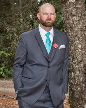 Man in gray three-piece suit with teal tie and coral boutonniere leaning against tree outdoors.