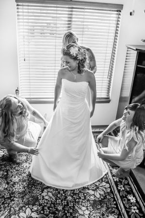 Bride in white wedding gown assisted by two bridesmaids in black and white photo near window with blinds.