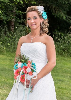 Bride in strapless white gown holding coral and turquoise bouquet with pearl accents, wearing matching hair flowers outdoors.
