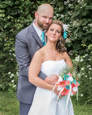 Bride in white strapless gown holding coral and turquoise bouquet stands with groom in gray suit outdoors.