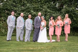 Wedding party posing outdoors, groomsmen in gray suits with teal ties and bridesmaids in coral dresses flanking bride and groom.