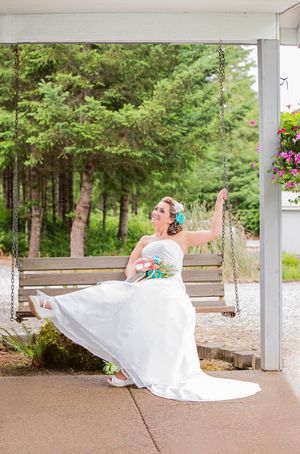 Bride in white gown sitting on a wooden porch swing, holding a colorful bouquet, with lush green trees in the background.
