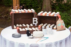 Wedding dessert table with cupcakes in wooden boxes, small cake with topper, and letter B decoration on white tablecloth.