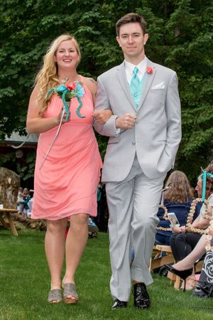 Bridesmaid in coral dress and groomsman in gray suit with teal tie walking down outdoor wedding aisle.