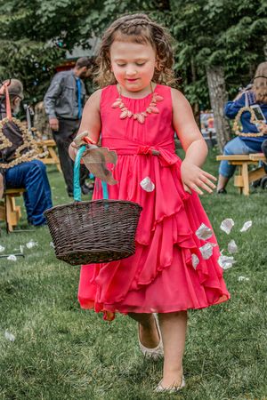 Young girl in red dress scattering flower petals from wicker basket at outdoor wedding ceremony.