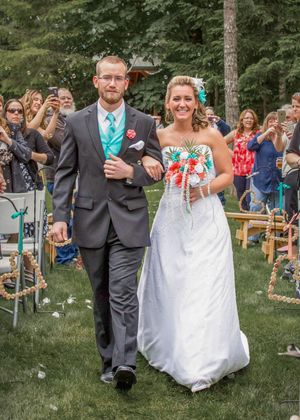 Newlywed couple walks down outdoor wedding aisle, groom in gray suit, bride in white gown holding coral and teal bouquet.