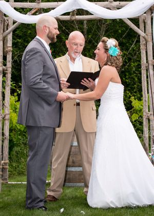 Bride and groom hold hands at outdoor wedding ceremony beneath rustic wooden arch with white draping.