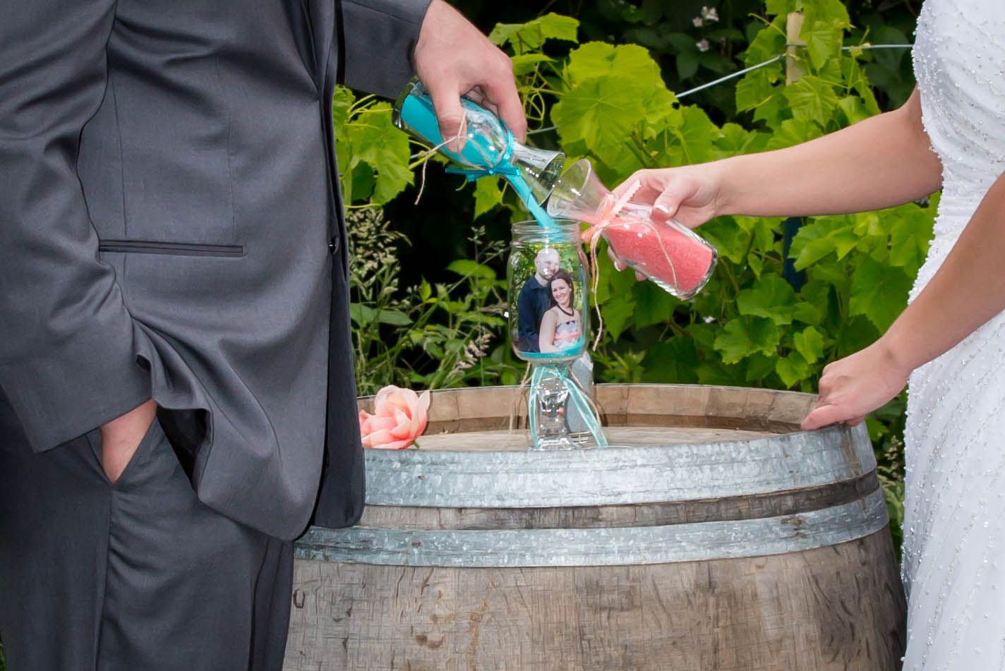 Bride and groom pouring colored sand into a vase during a unity ceremony on a wooden barrel at a vineyard wedding.