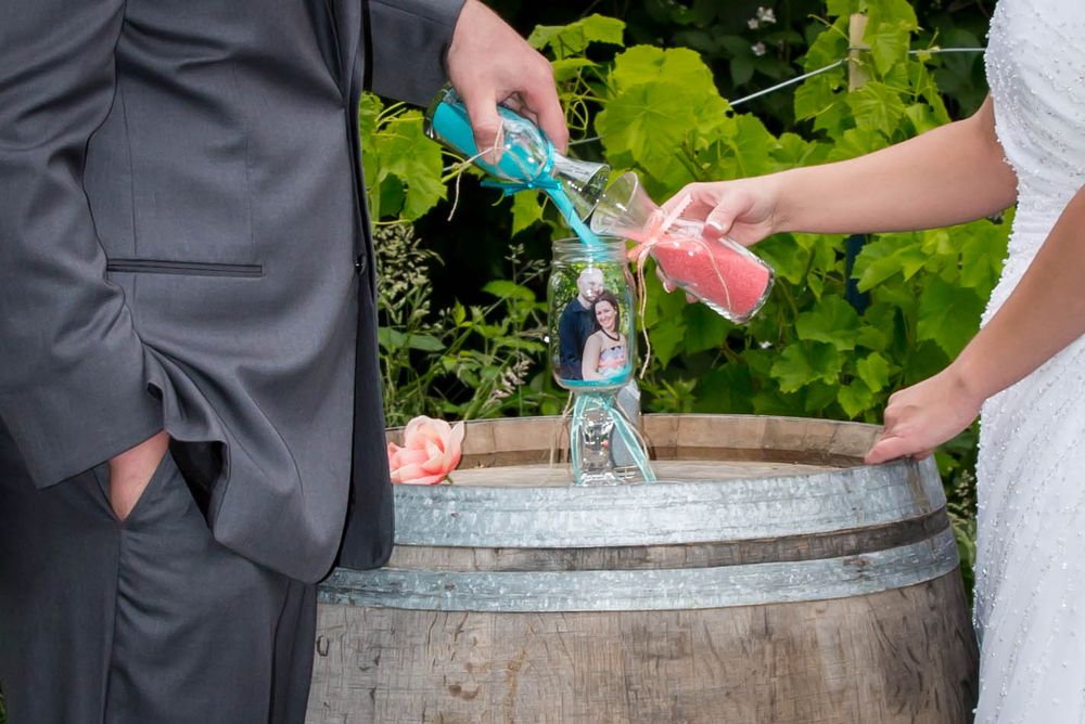 Bride and groom pouring colored sand into a vase during a unity ceremony on a wooden barrel at a vineyard wedding.
