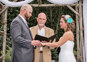 Bride and groom exchanging vows under rustic wooden arch with officiant during outdoor wedding ceremony.