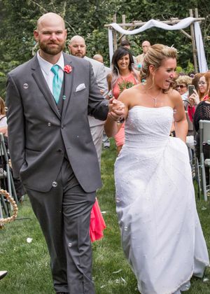 Newlywed couple holding hands walking down outdoor wedding aisle, groom in gray suit with teal tie, bride in white strapless gown.
