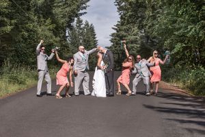 Wedding party celebrates on a tree-lined road, bride and groom kissing while groomsmen and bridesmaids in coral dresses cheer.