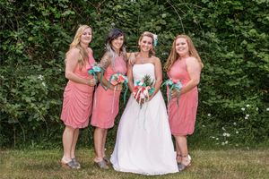 Bride in white gown with three bridesmaids in coral dresses holding bouquets, posing outdoors against a green hedge backdrop.