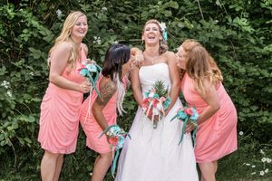 Bride in white dress laughs with three bridesmaids in coral pink dresses holding turquoise bouquets outdoors.