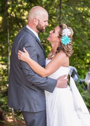 A bride and groom share a romantic embrace outdoors, she wearing a white lace gown with teal flower hair accessory.