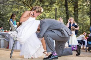 Groom kneeling to put shoe on bride in white wedding dress outdoors at rustic woodland wedding reception.