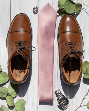 Brown leather dress shoes arranged with a pink tie and watch on white wooden background.
