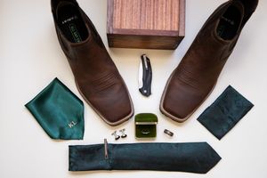 Brown leather square-toe boots with green tie, pocket square, cufflinks, ring box, and pocket knife on white background.