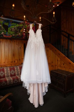 Elegant white lace wedding dress hanging on chandelier in rustic wood-paneled venue with staircase.