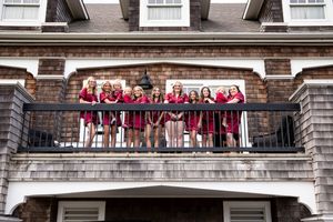 Group of women in red dresses posing on a balcony of a shingle-style building for a celebration photo.