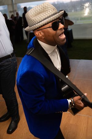 Man in blue velvet blazer, fedora hat, and sunglasses laughing at an outdoor event.