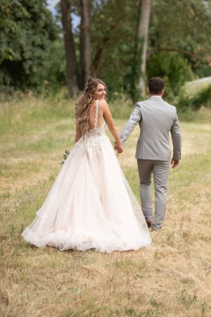 Newlyweds walking hand in hand through a grassy field in elegant wedding attire.