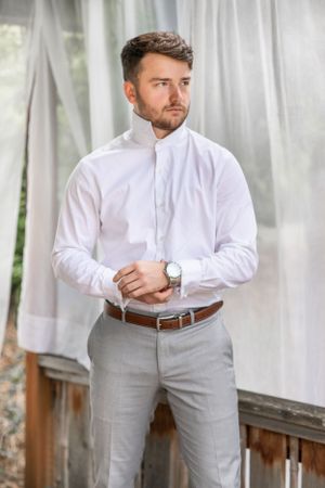 Groom in formal white shirt and grey pants adjusts cufflink against white curtain backdrop.