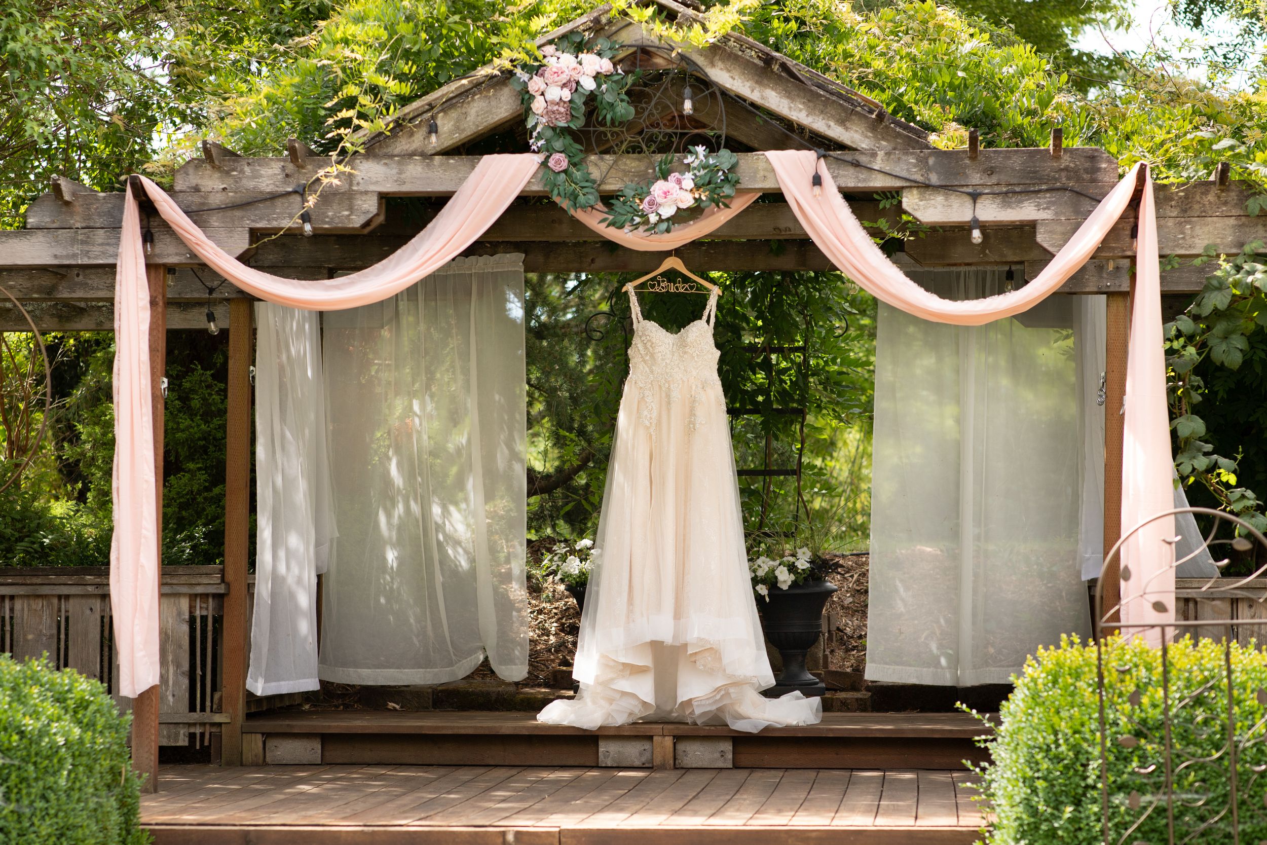 A white wedding dress hanging from a wooden pergola draped with sheer fabric.