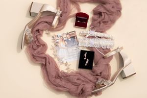 Flat lay of wedding accessories including shoes, perfume, and jewelry on a pink background.