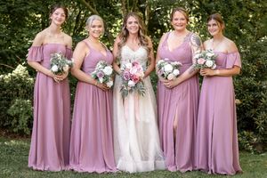 Bride in white gown with four bridesmaids in dusty rose dresses holding bouquets outdoors.