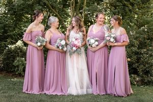 Bride in white gown with four bridesmaids in dusty rose dresses holding bouquets outdoors.