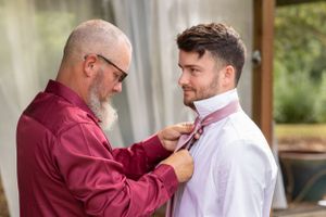 An older man in a burgundy shirt adjusting the collar of a younger man in a white shirt.