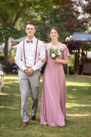 Wedding party members walking arm in arm outdoors, with the bridesmaid wearing a dusty rose gown and holding a bouquet.