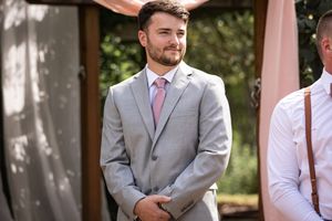 Groom in gray suit and pink tie smiling at wedding ceremony outdoors.
