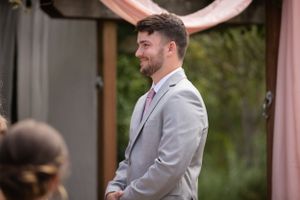 Side profile of a wedding guest in a light grey suit smiling during an outdoor ceremony.