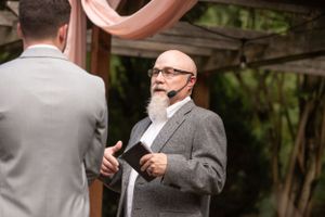 An officiant in a grey suit speaking during an outdoor wedding ceremony.